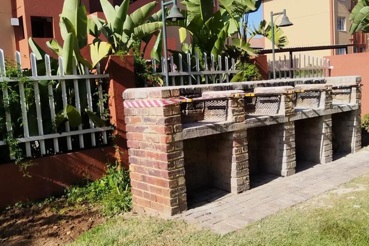 Communal outdoor area featuring a built-in brick braai station, paved walkway, and metal perimeter fencing set within a landscaped garden.