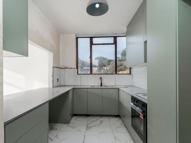 A modern kitchen in the final stages of renovation, featuring sage green cabinetry, stone countertops, and marble-look floor tiles, awaiting final wall finishes.
