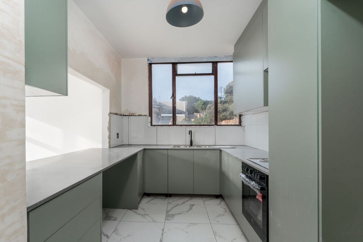A modern kitchen in the final stages of renovation, featuring sage green cabinetry, stone countertops, and marble-look floor tiles, awaiting final wall finishes.