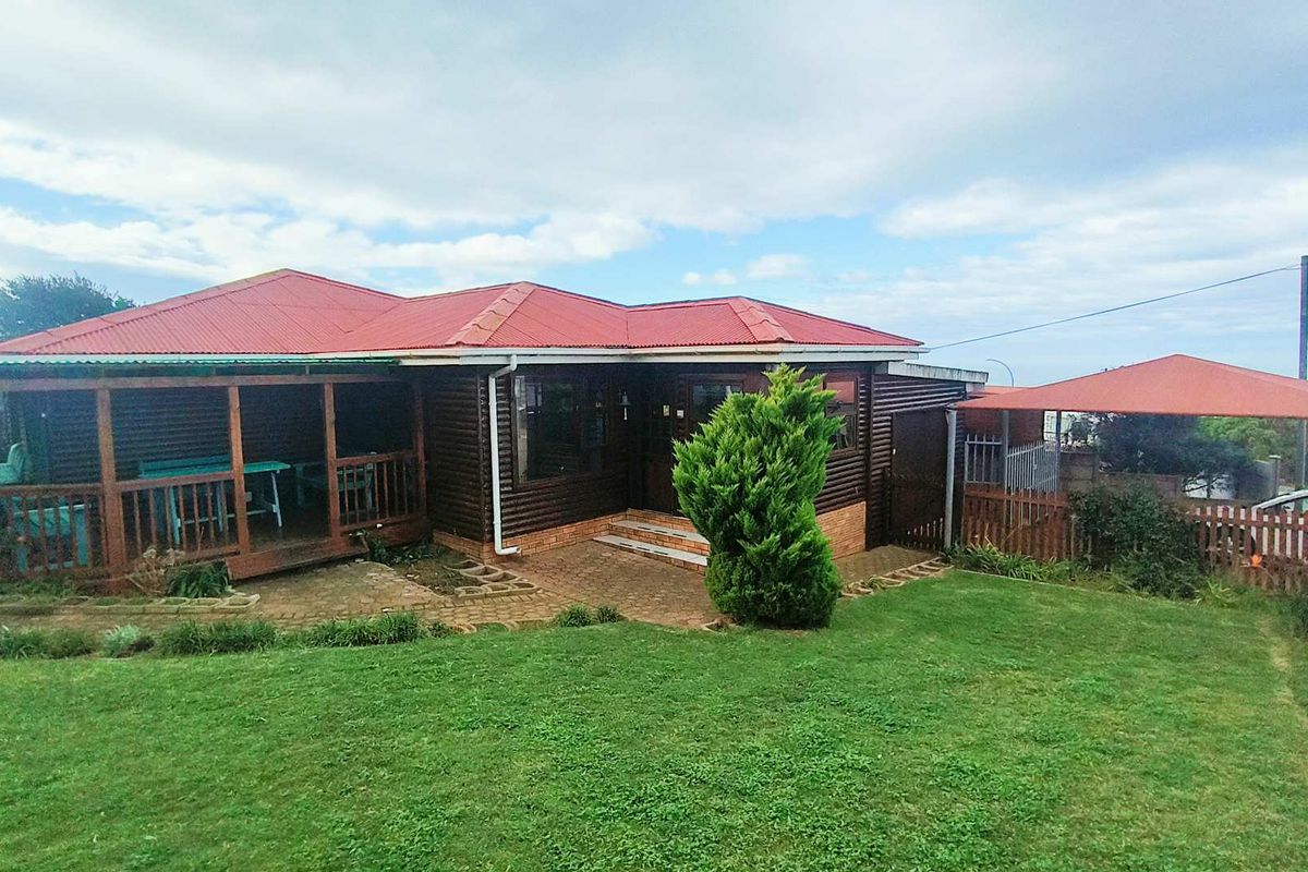 A charming log-style home featuring a covered timber deck, red corrugated metal roof, and a well-maintained garden with paved entrance area.