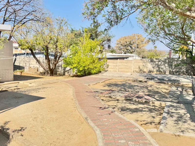 A spacious rear garden featuring mature trees, a brick paved pathway, and a precast concrete boundary wall, offering potential for landscaping improvements.