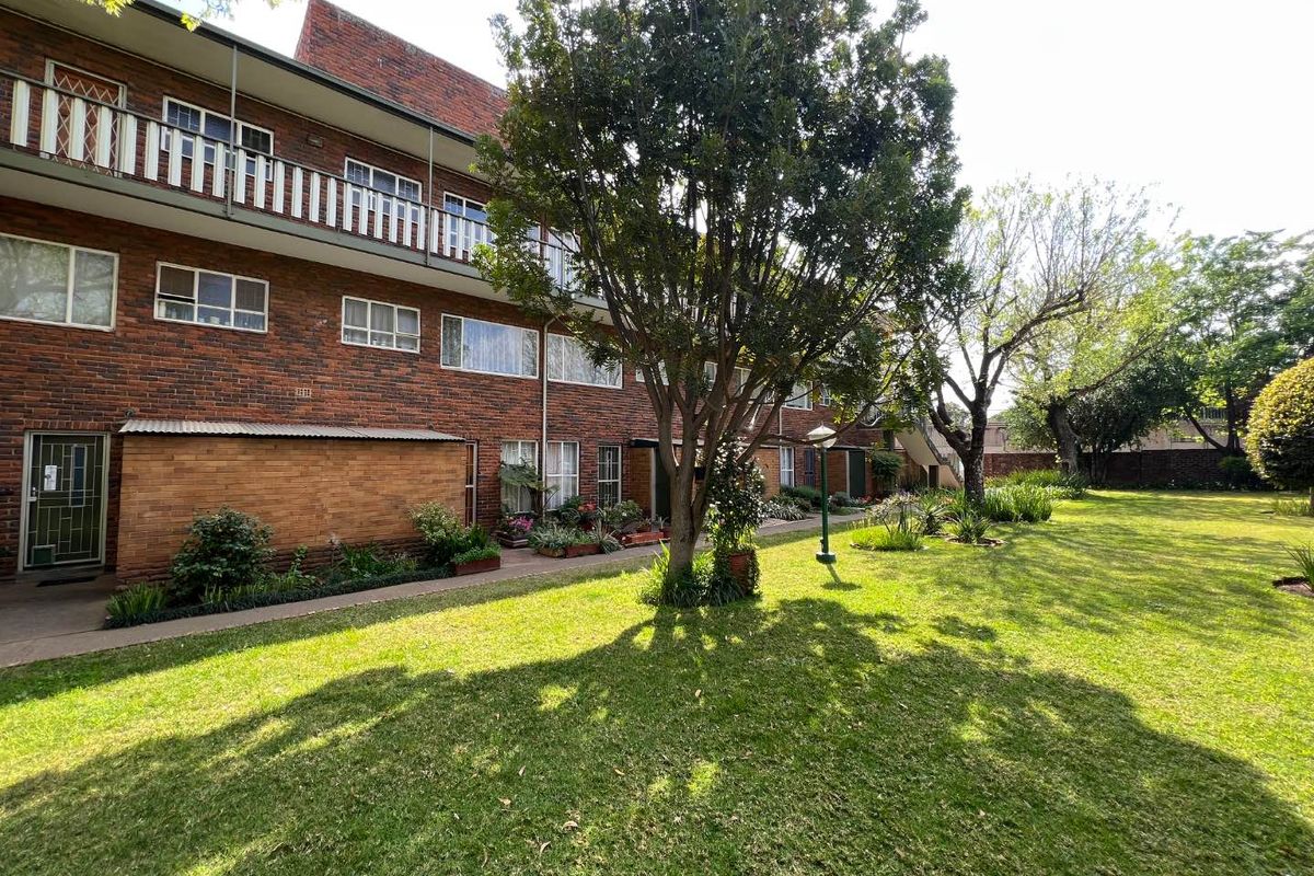 A view of the apartment complex exterior featuring a well-maintained communal garden, mature trees, and brick facade with balcony access.