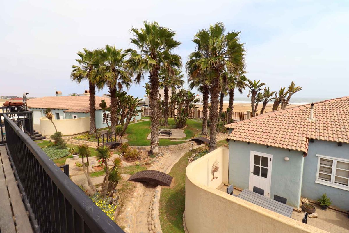 A well-maintained communal garden area featuring mature palm trees, stone pathways, and wooden footbridges, set between residential units with views toward the coast.