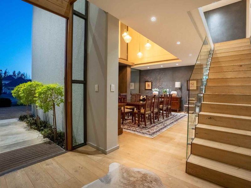 Elegant entrance foyer featuring a timber staircase with glass balustrade, leading into a spacious dining area with warm recessed lighting.