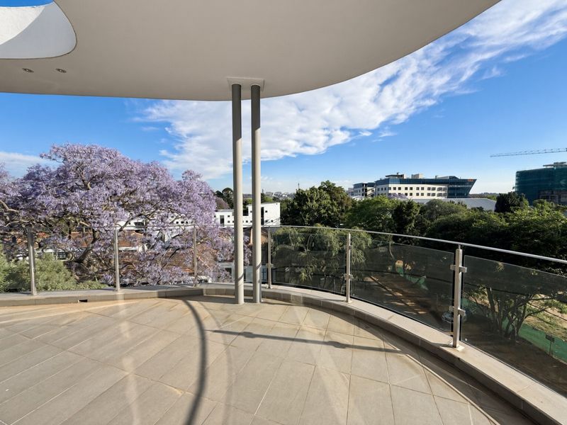 A spacious, modern balcony featuring tiled flooring, a sleek glass balustrade, and a covered area with recessed lighting, offering expansive neighborhood views.