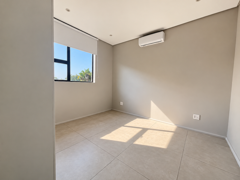 A bright, empty bedroom featuring neutral tiled flooring, recessed ceiling lights, and a wall-mounted air conditioning unit with a window view.