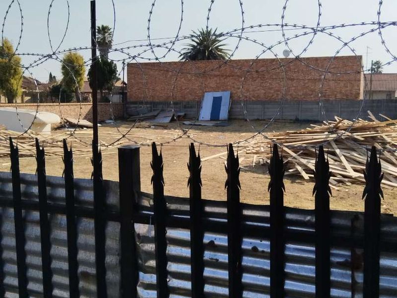 Vacant residential land secured with metal palisade fencing and razor wire, featuring a brick boundary wall and scattered construction debris on site.