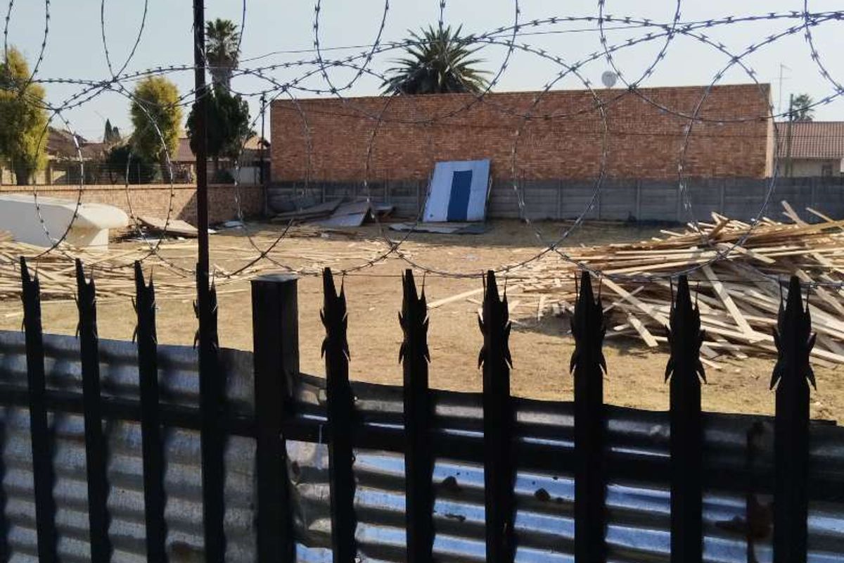 Vacant residential land secured with metal palisade fencing and razor wire, featuring a brick boundary wall and scattered construction debris on site.