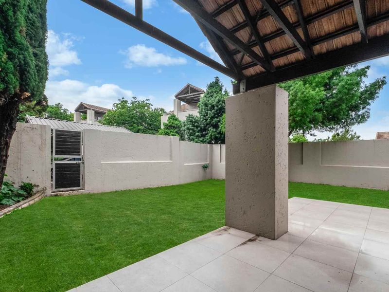 A covered patio area featuring tiled flooring and an exposed timber roof structure, overlooking a private garden with perimeter walling.