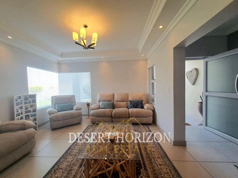 A well-lit living room featuring a tray ceiling, recessed lighting, and neutral tiled flooring with large windows fitted with venetian blinds.