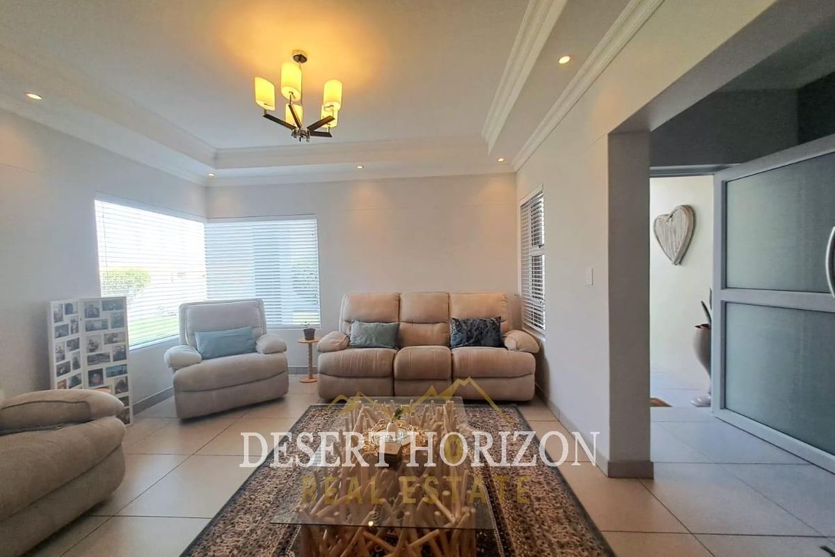 A well-lit living room featuring a tray ceiling, recessed lighting, and neutral tiled flooring with large windows fitted with venetian blinds.