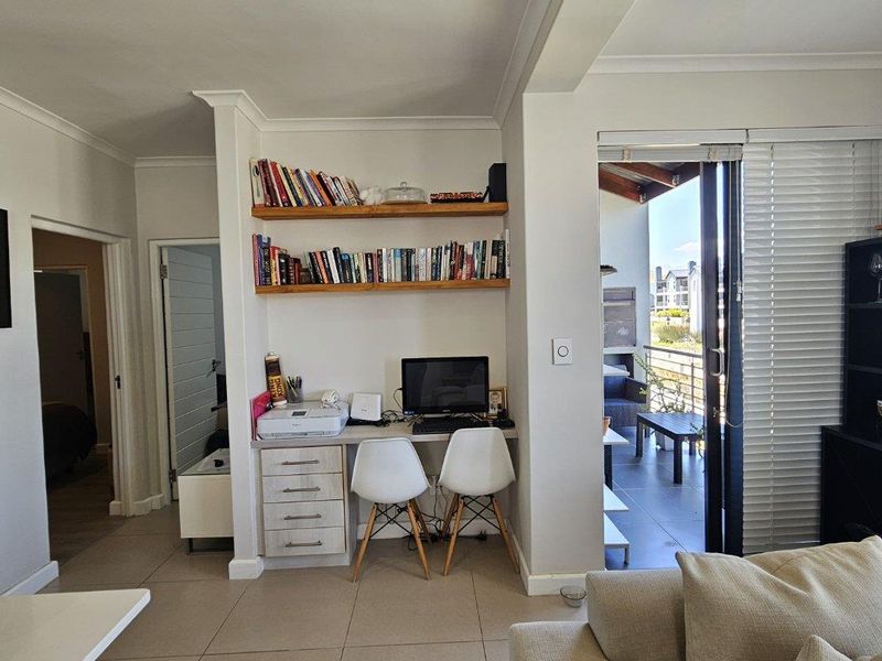 A bright living area featuring a functional built-in study nook with shelving, tiled floors, and direct access to an outdoor balcony through sliding doors.