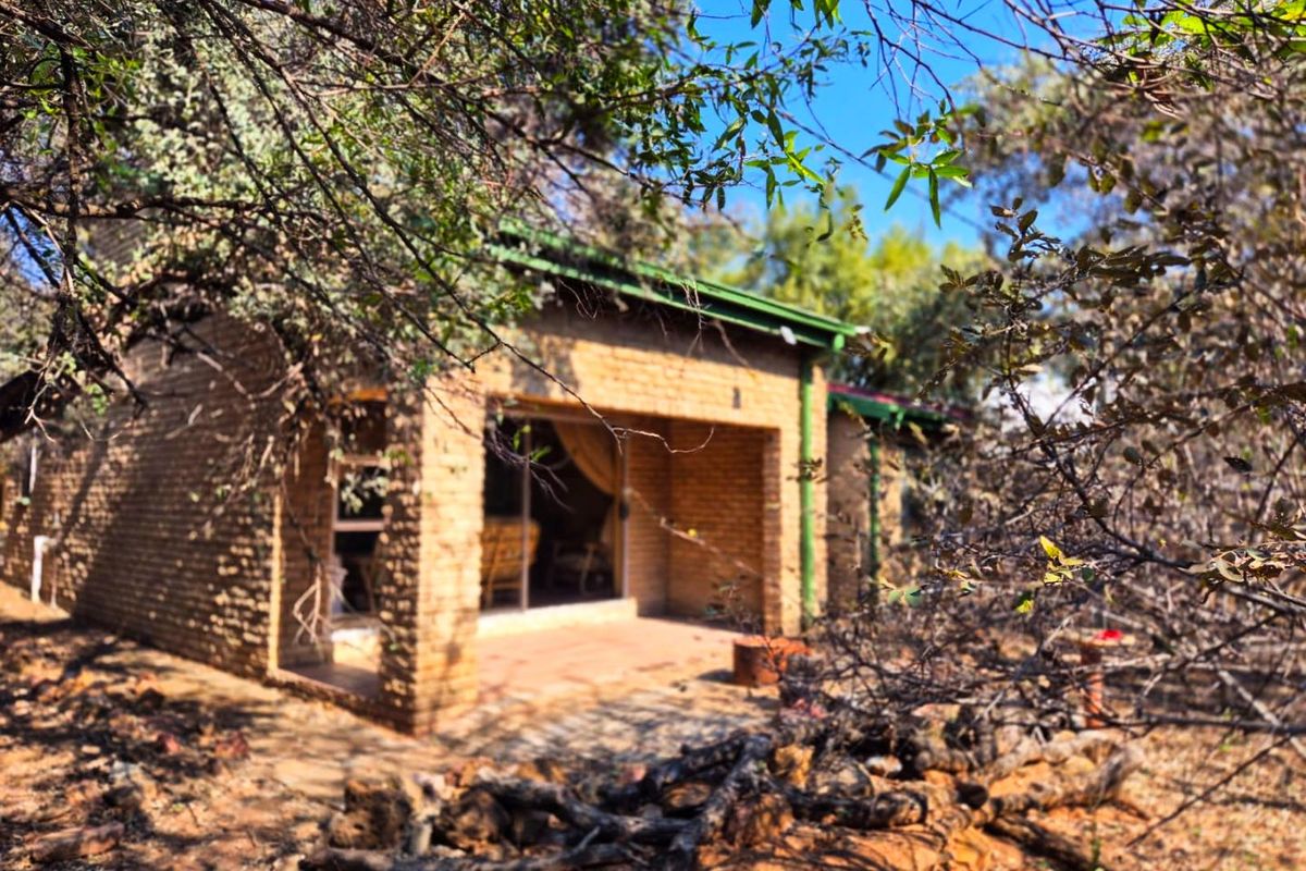 A view of the rear exterior of a brick house featuring a covered patio and sliding glass doors, surrounded by natural bushveld vegetation.