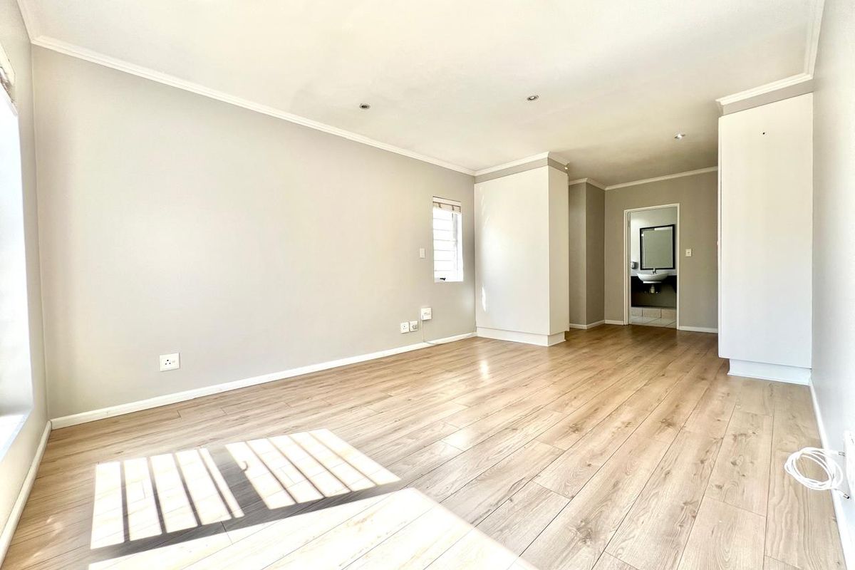 A bright, empty bedroom featuring light wood-look laminate flooring, built-in cupboards, and recessed downlights, with an adjoining bathroom visible through the doorway.