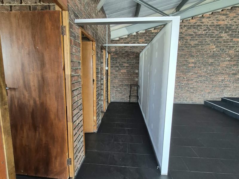 An open-plan office space featuring exposed brick walls, steel roof trusses, and dark tiled flooring with an unfinished partition wall.