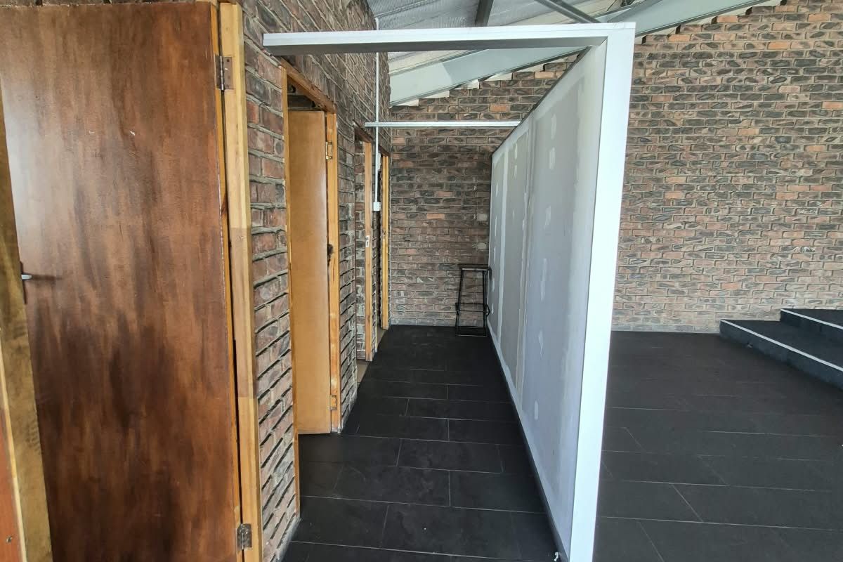 An open-plan office space featuring exposed brick walls, steel roof trusses, and dark tiled flooring with an unfinished partition wall.