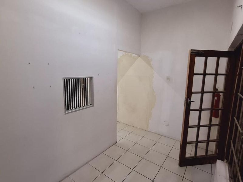 An empty office space featuring tiled flooring, a security window, and a timber-framed glass door, currently requiring wall finishing and cosmetic repairs.