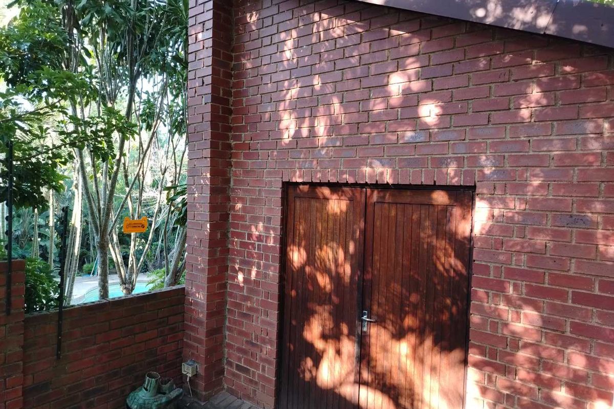 A view of a red brick exterior wall featuring double wooden doors, situated in a garden setting with mature trees.
