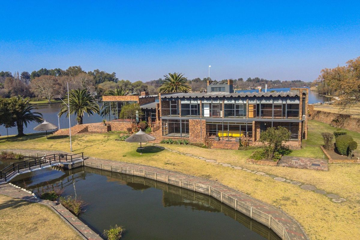 Aerial view of a large brick waterfront residence featuring extensive glass windows, a private jetty, and expansive landscaped grounds.