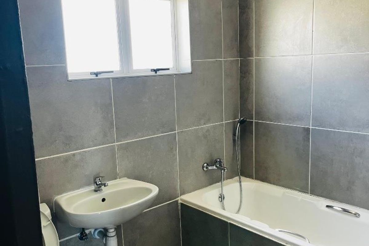 A compact bathroom featuring full-height grey wall tiling, a built-in bathtub with shower mixer, a wall-mounted basin, and a standard toilet.