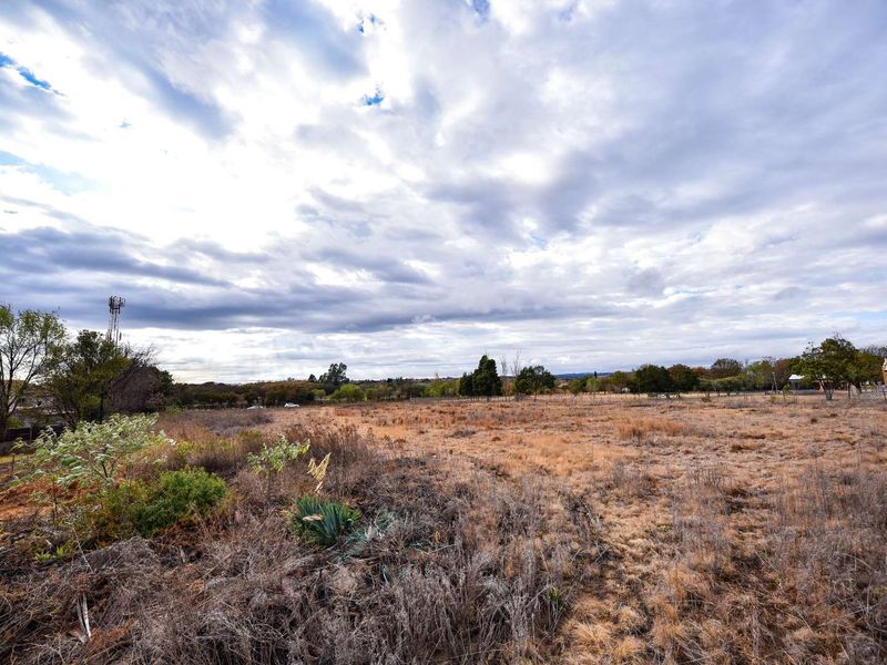 A wide-angle view of a vacant residential plot featuring dry grass, natural vegetation, and perimeter wire fencing under a cloudy sky.