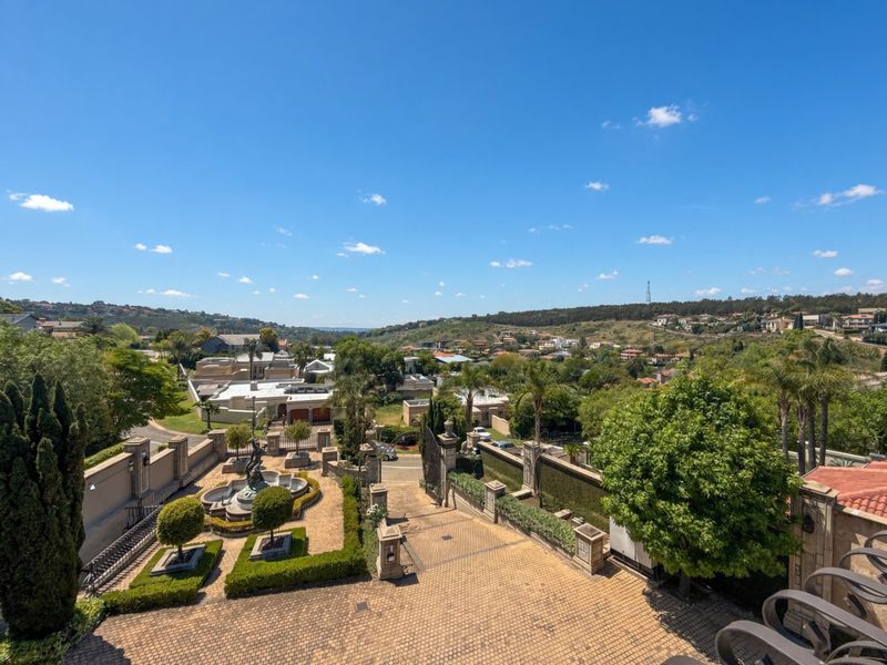 Elevated view overlooking a well-maintained estate entrance, featuring a paved driveway, stone boundary walls, and an ornamental fountain within a landscaped garden setting.