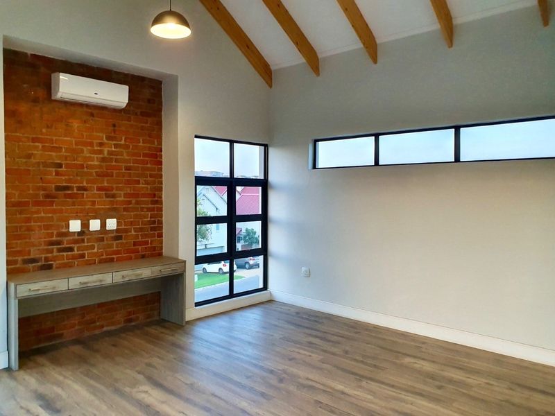 A bright, modern living space featuring a vaulted ceiling with exposed timber beams, an exposed brick feature wall, built-in desk, and laminate flooring.
