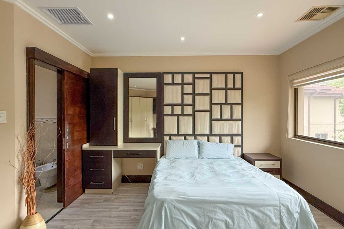 A well-maintained bedroom featuring built-in cabinetry, a custom headboard, recessed ceiling lighting, and an en-suite bathroom accessible via a sliding wooden door.