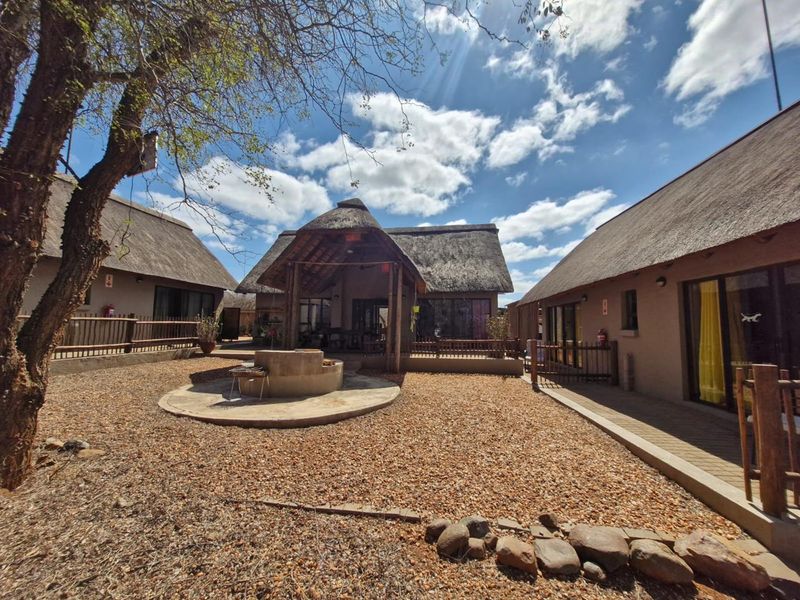A spacious rear courtyard featuring a central built-in braai area, surrounded by multiple structures with traditional thatch roofing and timber decking.