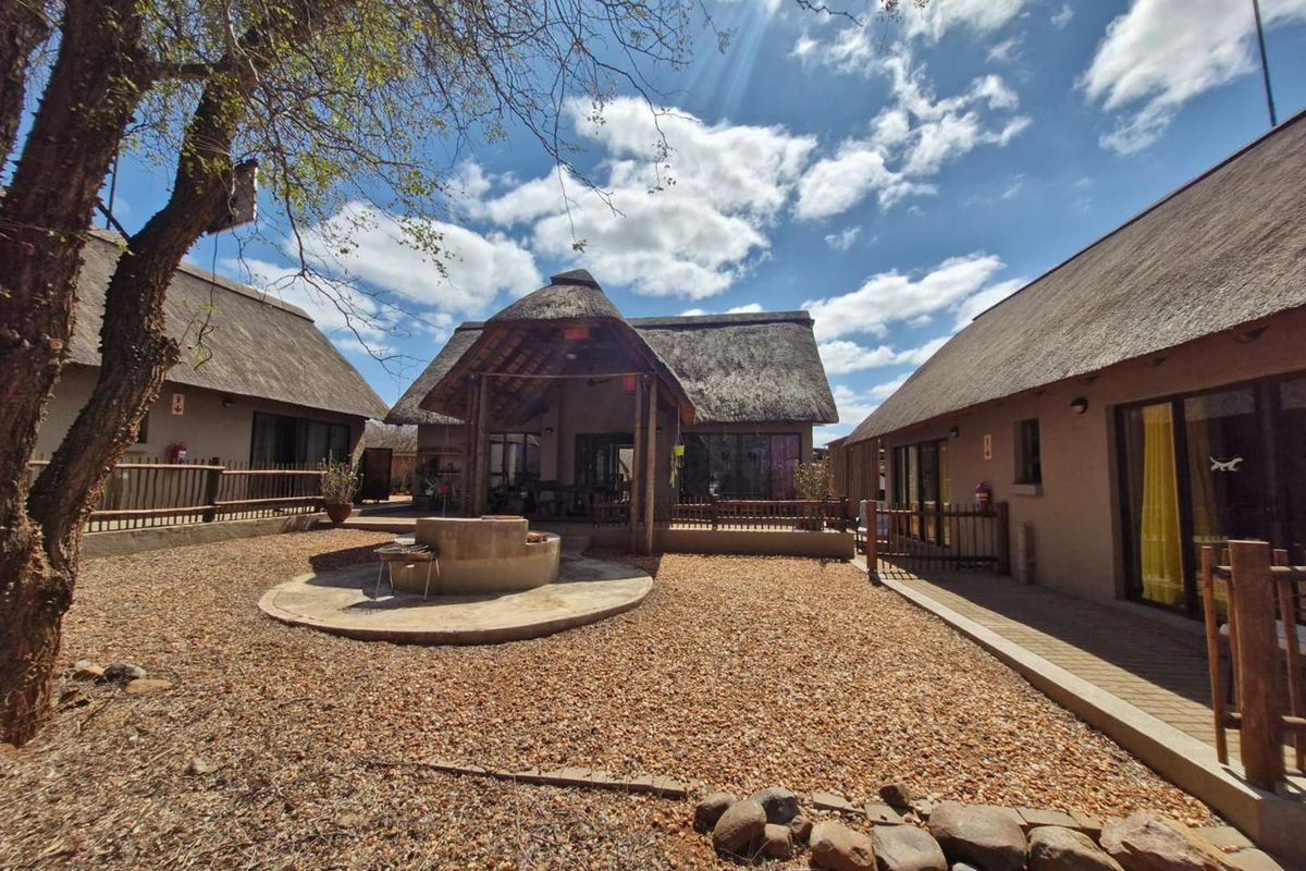 A spacious rear courtyard featuring a central built-in braai area, surrounded by multiple structures with traditional thatch roofing and timber decking.