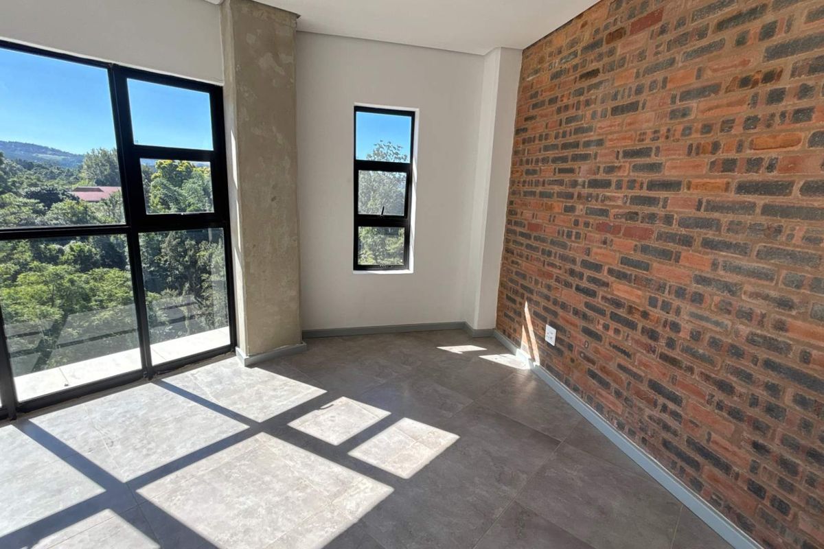 A bright, empty bedroom featuring a striking exposed brick feature wall, large floor-to-ceiling windows, and modern grey floor tiles.