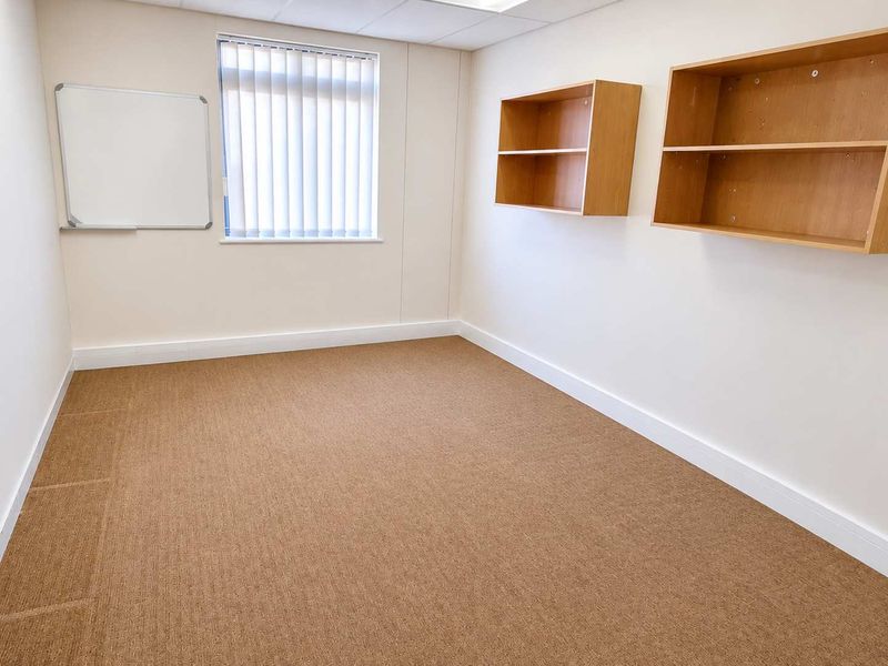 A clean, empty office space featuring neutral walls, carpeted flooring, vertical blinds, and built-in wall shelving units.