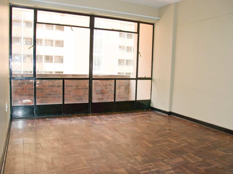 An empty living room featuring original parquet flooring and large metal-framed windows with views of the adjacent building.
