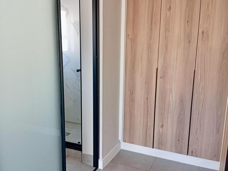 Modern bedroom area featuring sleek wood-finish built-in cupboards, neutral floor tiles, and a contemporary sliding glass door leading to an en-suite bathroom.