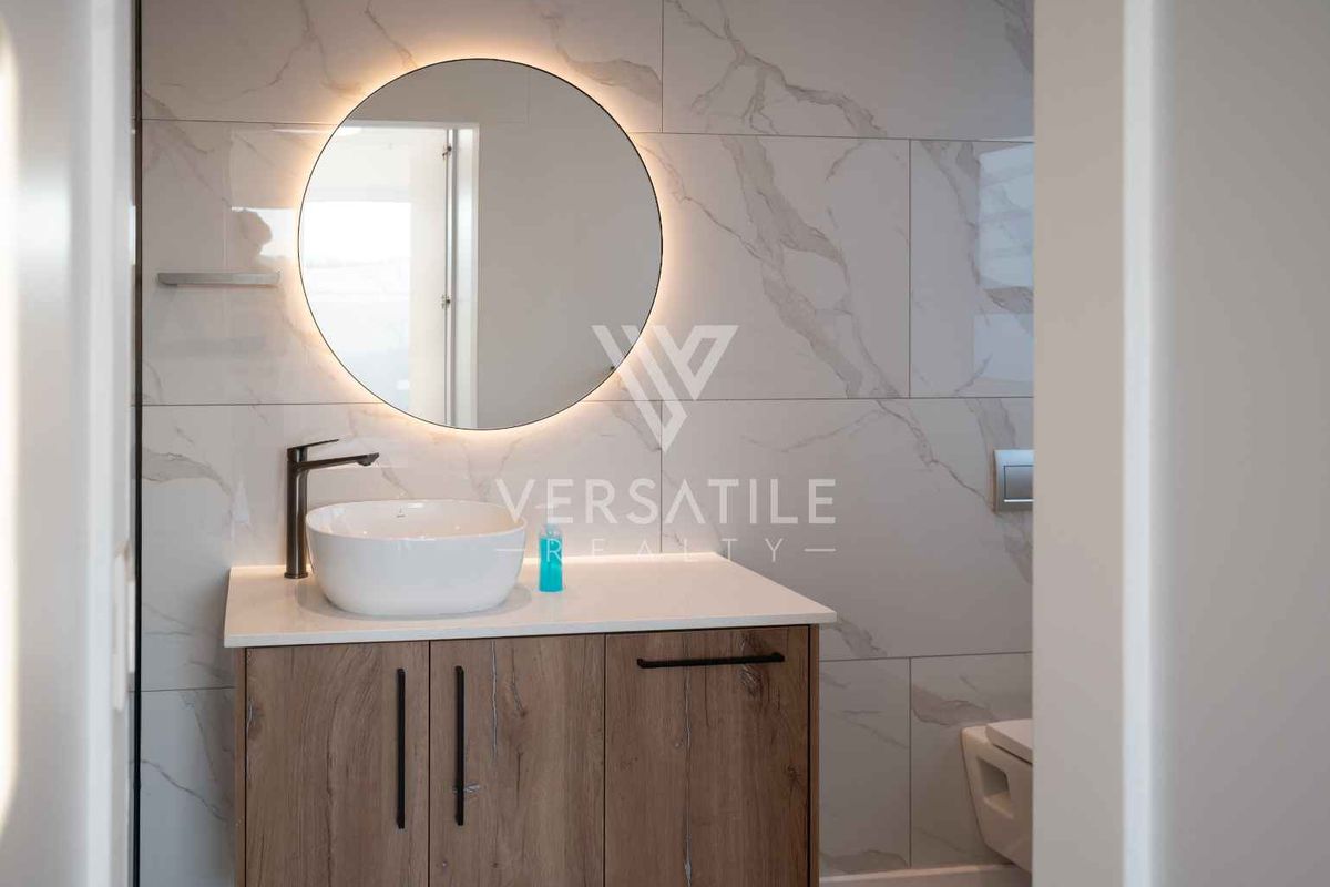 Modern bathroom featuring a wood-finish vanity, countertop basin, backlit circular mirror, and elegant marble-look wall tiles.