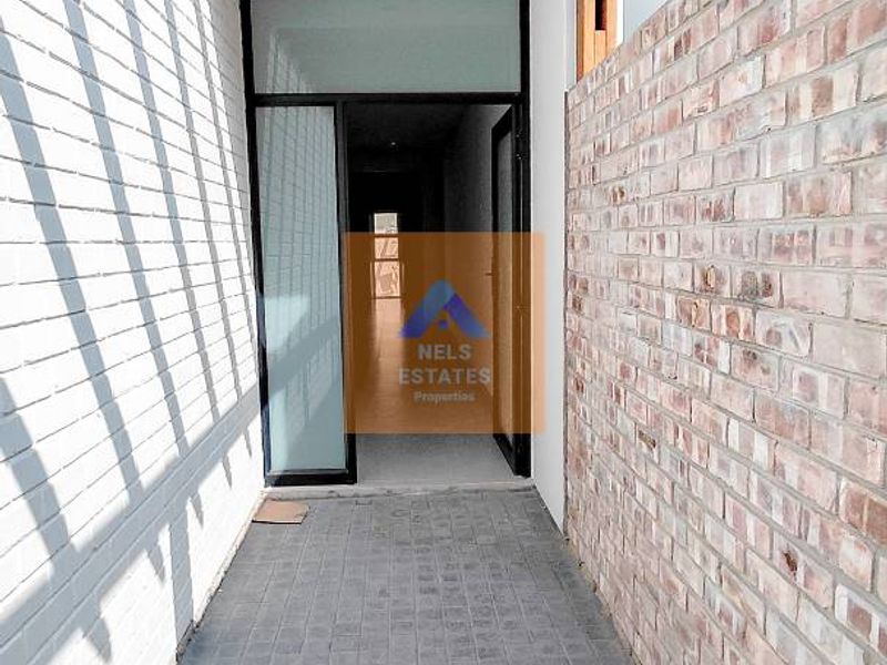 A modern outdoor walkway or patio area featuring a timber pergola, exposed brickwork, and paved flooring leading to a glass entrance door.