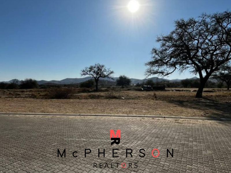 A vacant commercial land plot featuring paved road access, kerbing, and utility connection points, set against a natural landscape with mountain views.