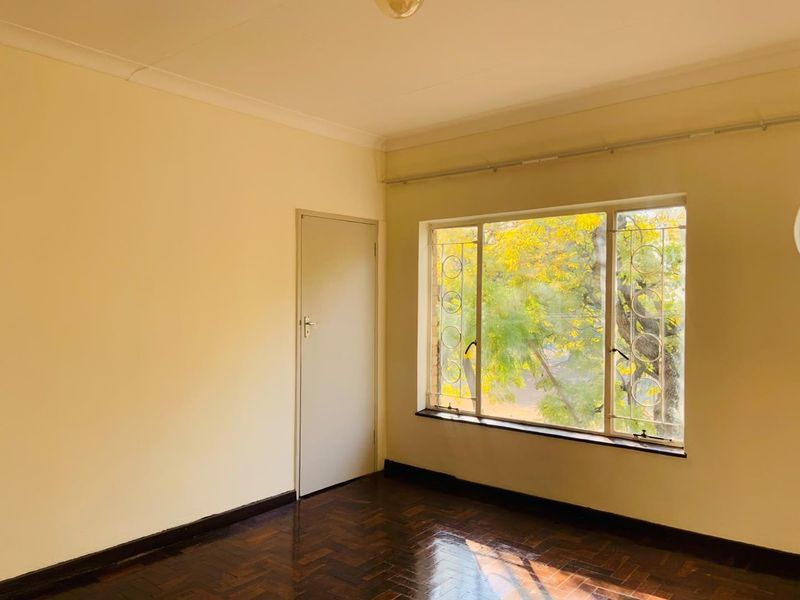 A bright, empty bedroom featuring polished parquet flooring, neutral walls, and large windows fitted with security bars.