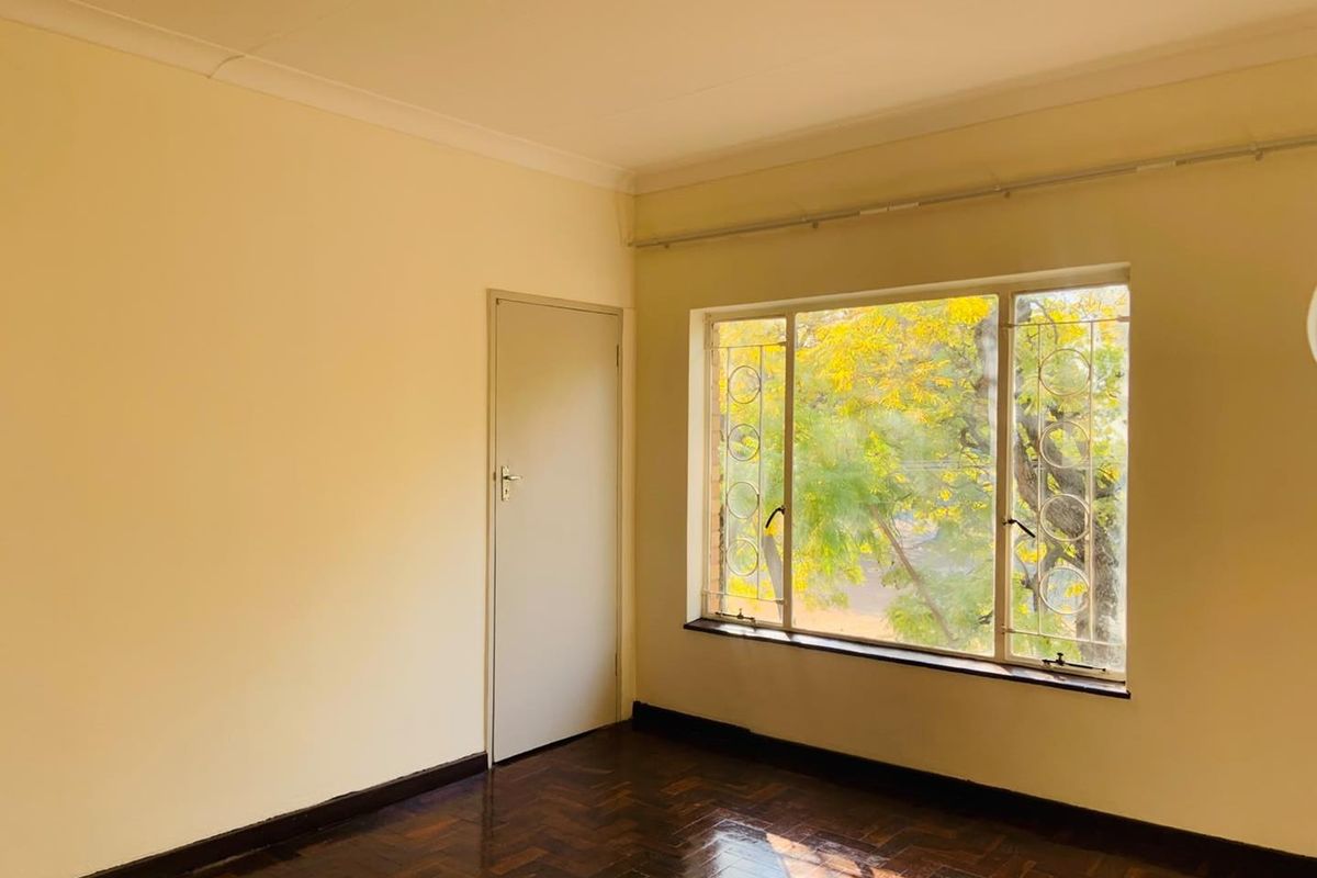 A bright, empty bedroom featuring polished parquet flooring, neutral walls, and large windows fitted with security bars.
