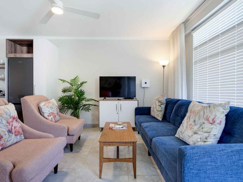 Open plan living area featuring tiled flooring, ceiling fan, and large windows with venetian blinds, seamlessly connected to a modern kitchen space.