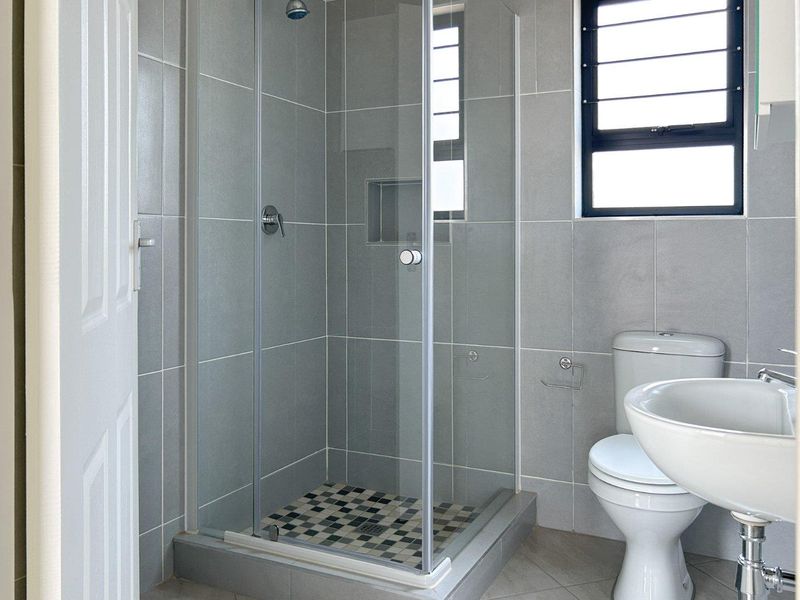 A clean, modern bathroom featuring a glass-enclosed shower with a built-in niche, pedestal basin, and toilet, finished with neutral grey wall tiles.