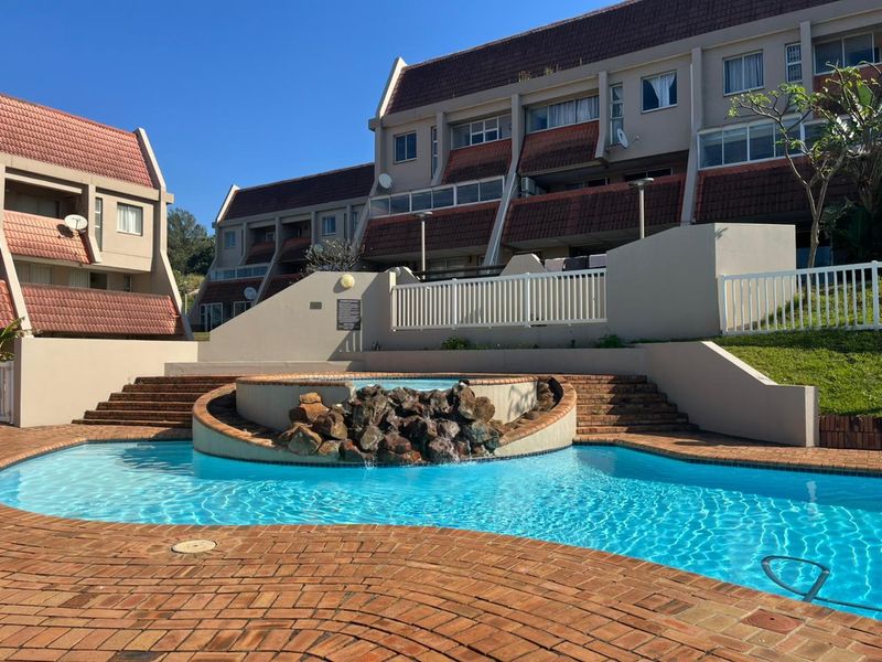 Communal swimming pool area featuring a decorative rock water feature, brick paving, and surrounding apartment complex architecture.