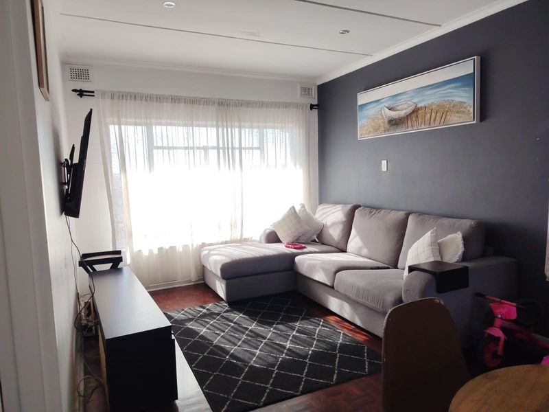 A comfortable living room featuring warm parquet flooring, a dark accent wall, and large windows that provide ample natural light.