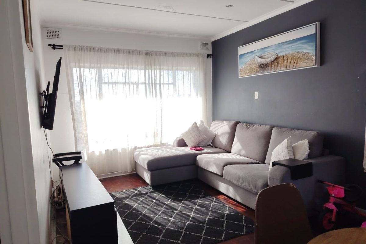 A comfortable living room featuring warm parquet flooring, a dark accent wall, and large windows that provide ample natural light.