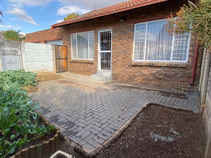 Rear exterior view of the townhouse featuring a brick facade, tiled roof, paved patio area, and a secure entrance with a metal security gate.