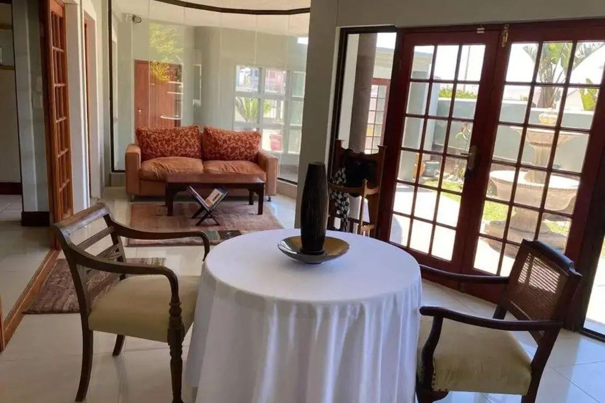 A bright living area featuring tiled flooring, large glass windows, and double wooden doors providing direct access to the outdoor garden space.
