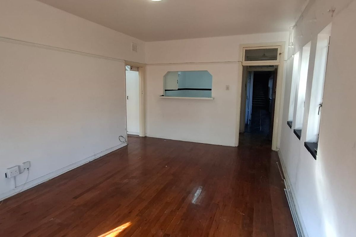 An empty living room featuring hardwood flooring, a serving hatch to the kitchen, and multiple windows, presenting a blank canvas for renovation.