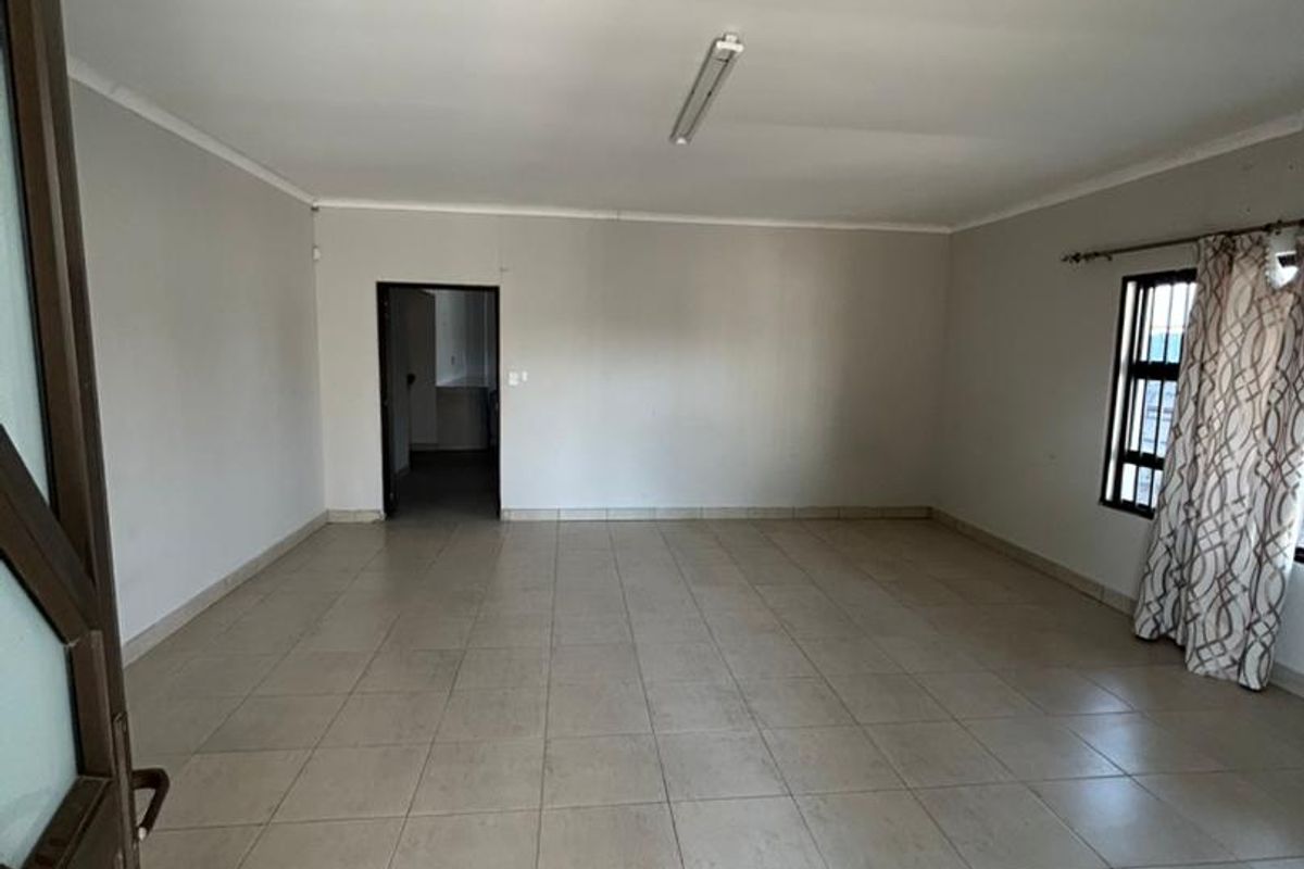 A spacious, empty living area featuring neutral ceramic floor tiles, a simple ceiling light fixture, and a window with security bars.