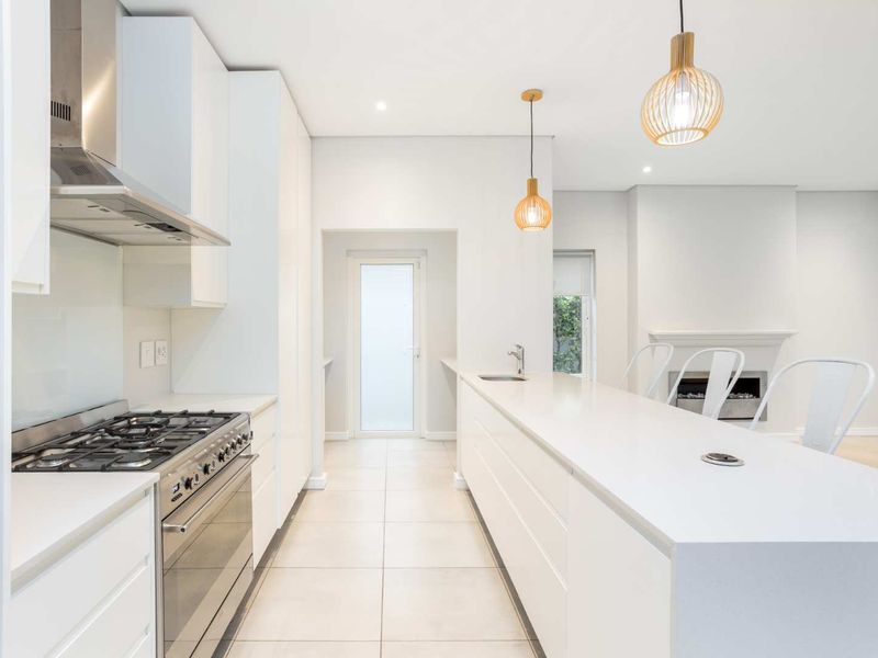 Modern, bright kitchen featuring sleek white cabinetry, stone countertops, a large island with integrated sink, and a gas stove, flowing into an open living area.