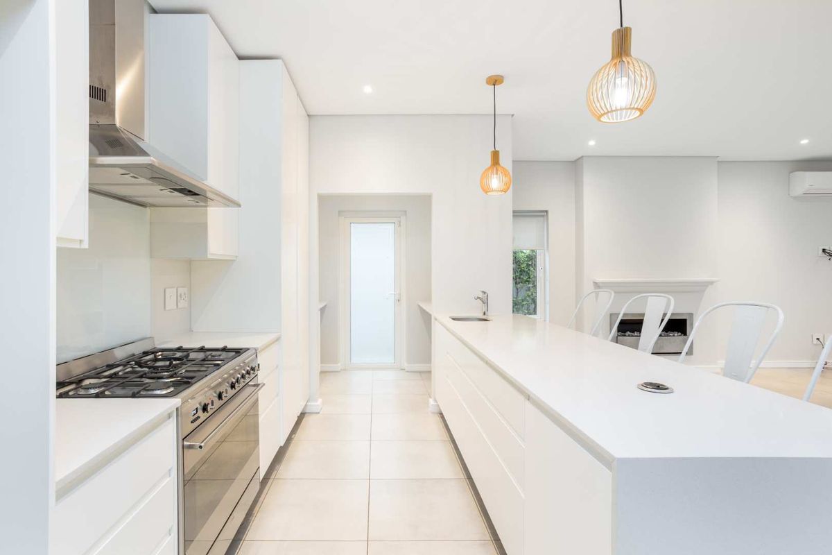 Modern, bright kitchen featuring sleek white cabinetry, stone countertops, a large island with integrated sink, and a gas stove, flowing into an open living area.